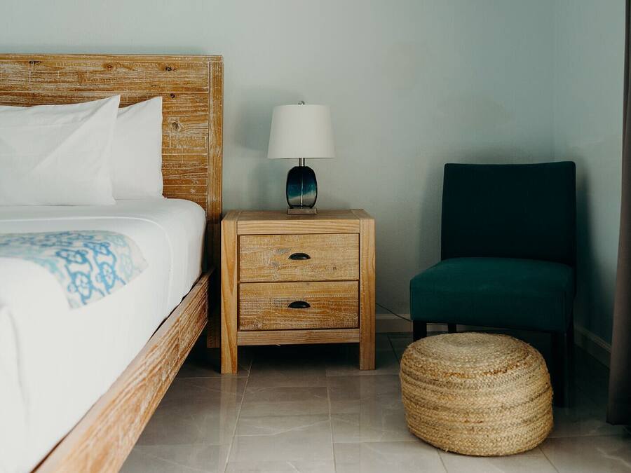 A hotel room with a wooden headboard bed, a nightstand with a lamp, a green chair, and a woven ottoman.