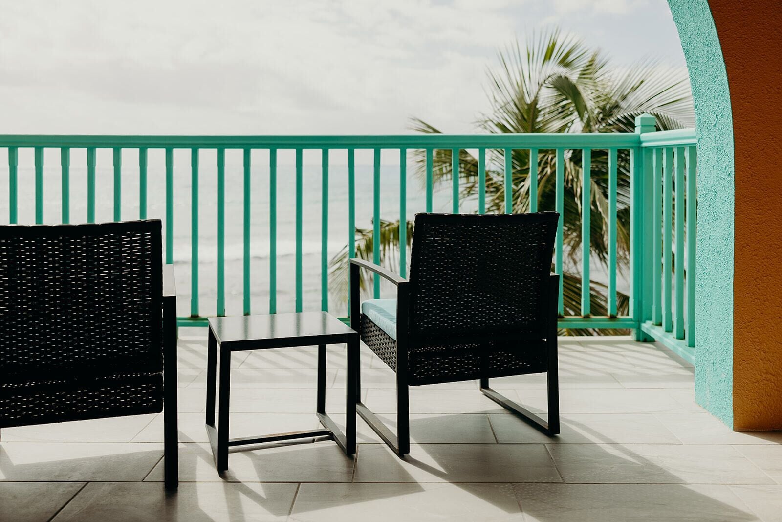 A balcony with two black chairs and a small table, a turquoise railing, and palm trees in the background.