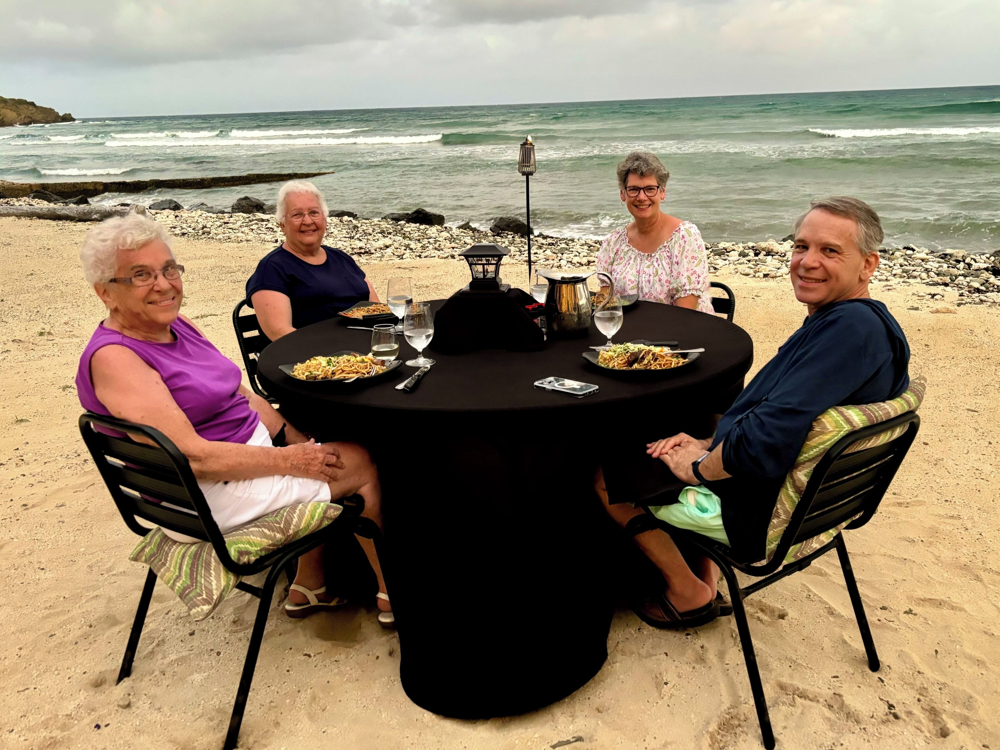 A group of people seated around a round table on a sandy beach with a view of the ocean.