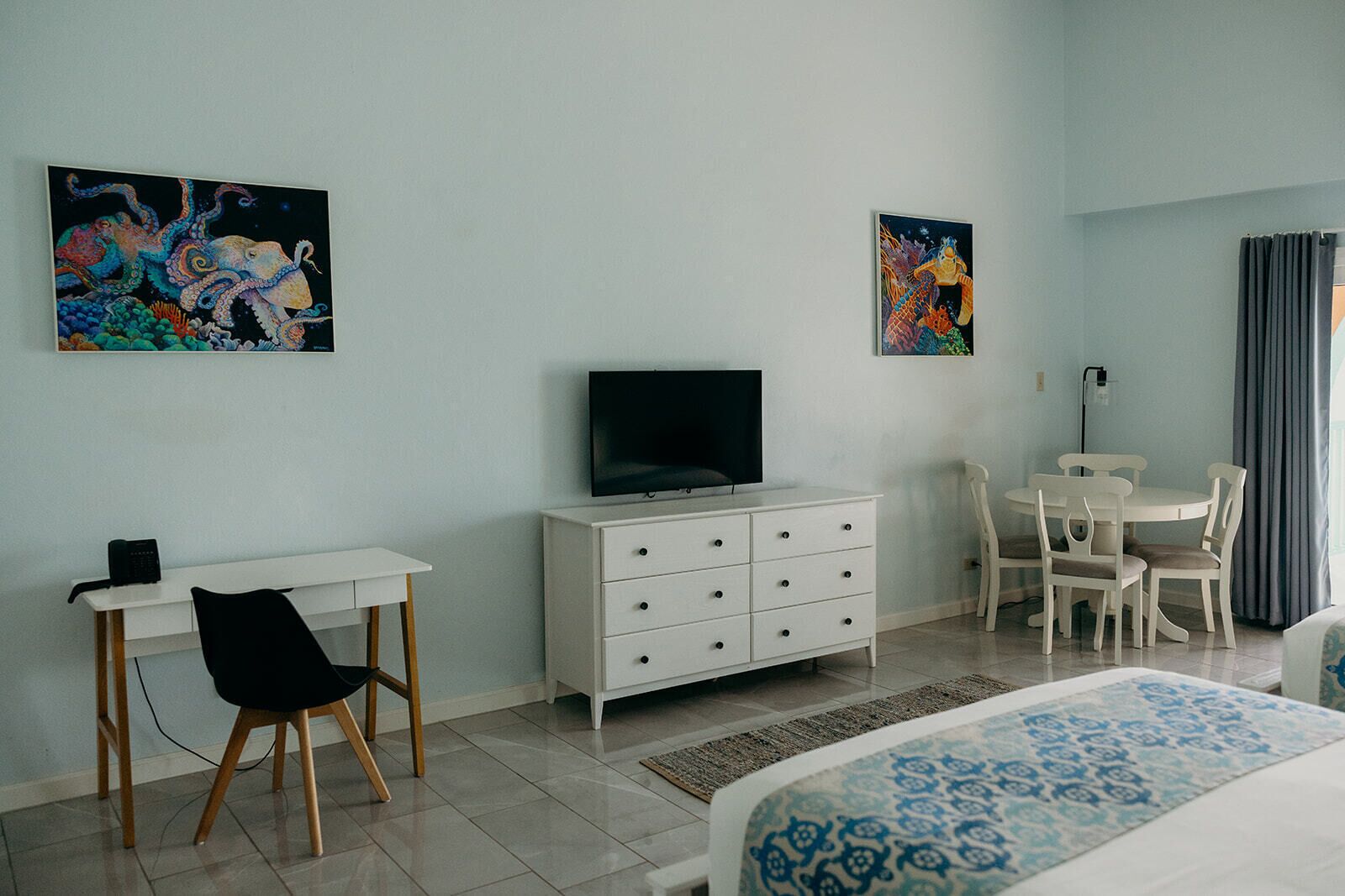 A hotel room with a bed, a desk with a chair, a television, a small table with chairs, and two paintings on the walls.