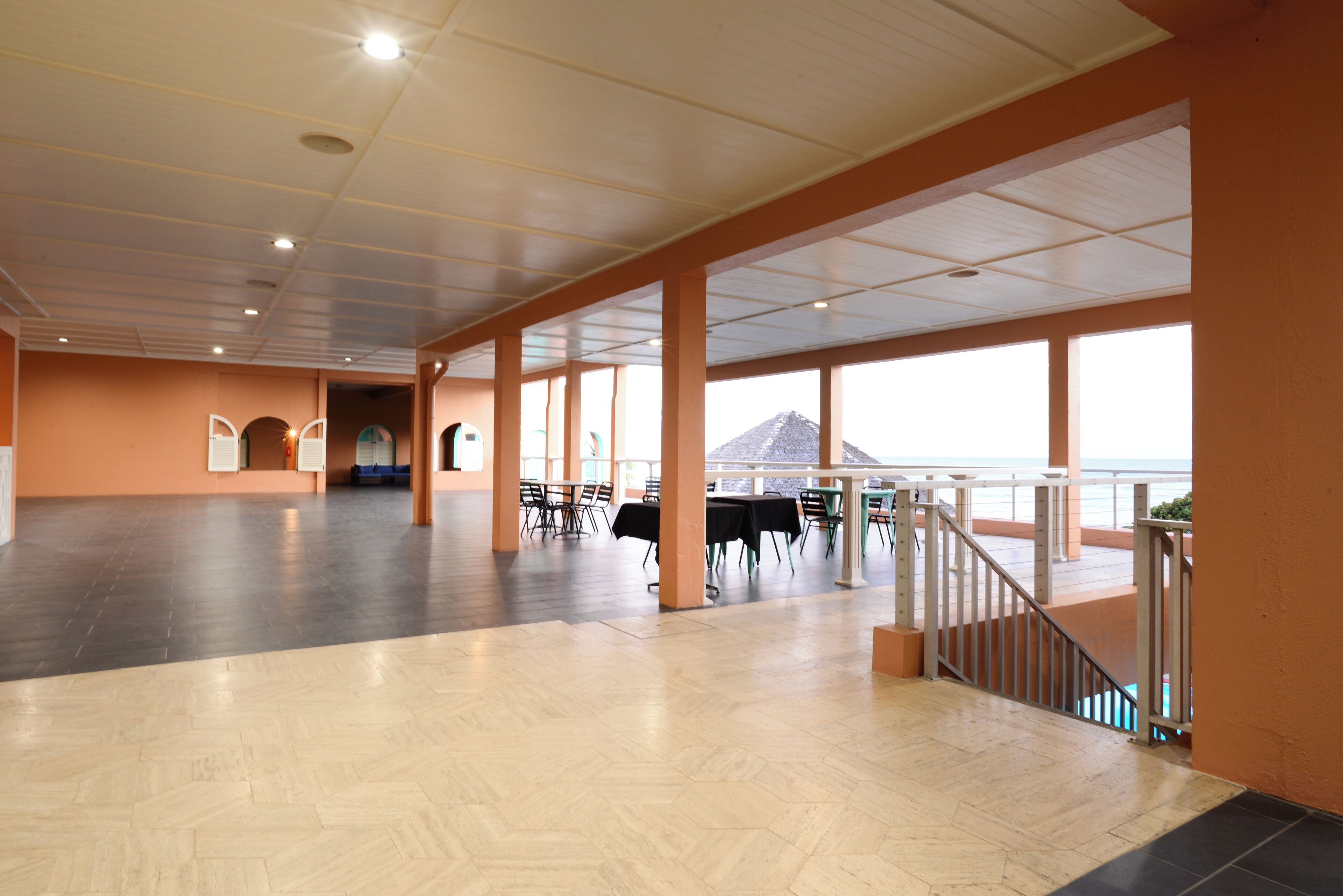 A spacious indoor area with a view of the sea, featuring a wooden floor, tables, chairs, and a staircase leading to an outdoor balcony.