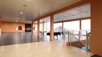A spacious indoor area with a view of the sea, featuring a wooden floor, tables, chairs, and a staircase leading to an outdoor balcony.