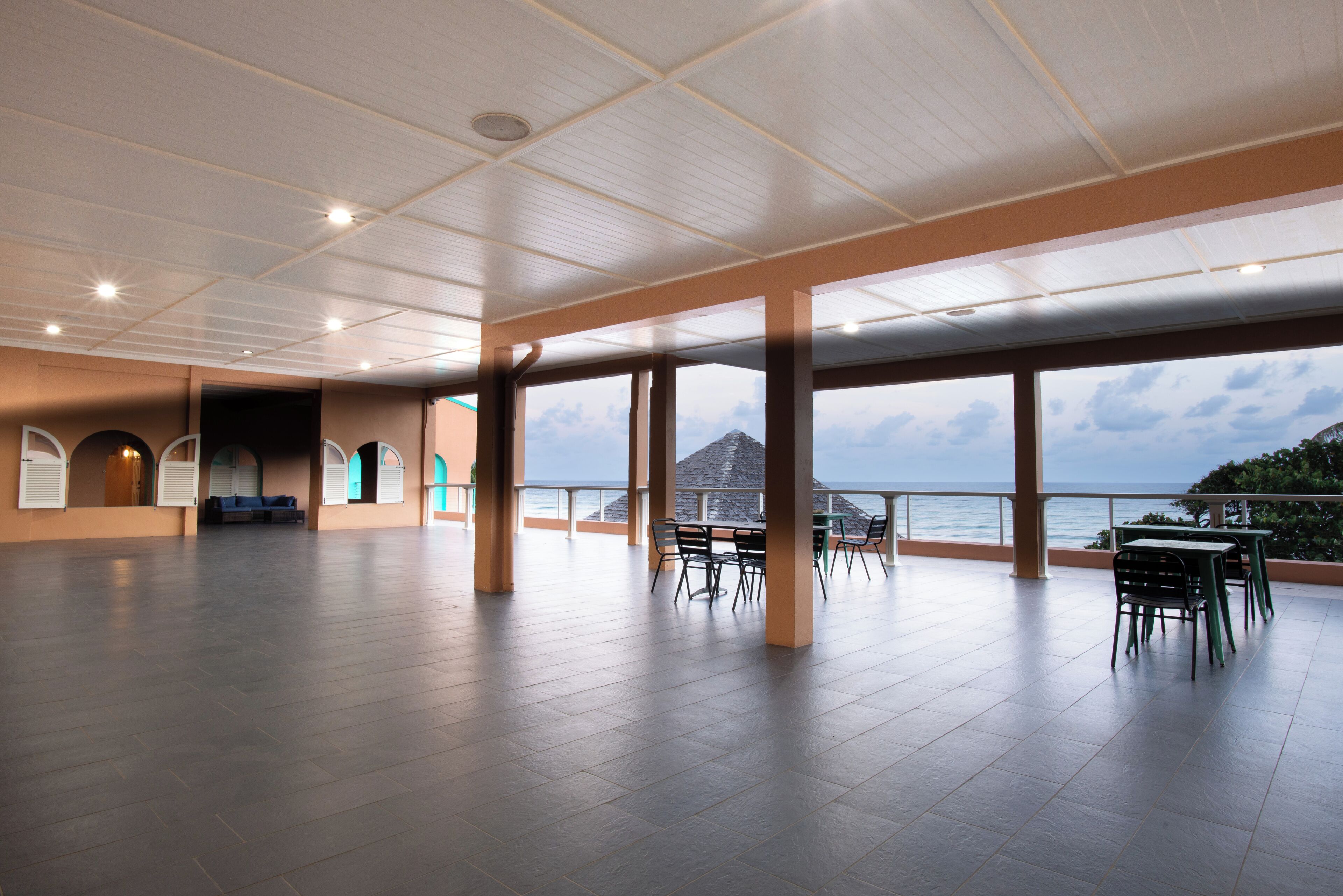 Un espacio interior amplio con vista al mar, que cuenta con un área abierta y techos altos, una zona de estar con mesas y sillas, y un balcón con barandilla.