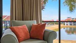 Un sillón verde con dos almohadas naranjas en una habitación con un ventanal grande que da a un cuerpo de agua y edificios.