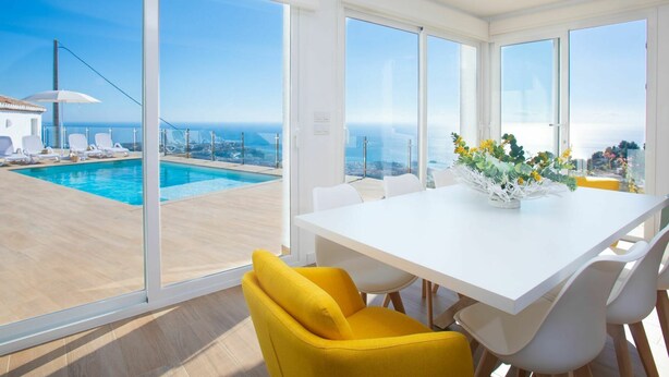 Un espace repas moderne avec une table blanche et des chaises jaunes, offrant une vue sur une piscine et l’océan.
