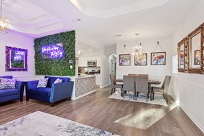 Una sala de estar moderna con un conjunto de sofá azul, una zona de comedor con una mesa grande y una cocina al fondo.