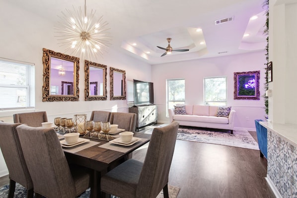 Un comedor moderno con una mesa de madera, sillas y objetos decorativos. Se aprecia un sofá y una televisión al fondo.