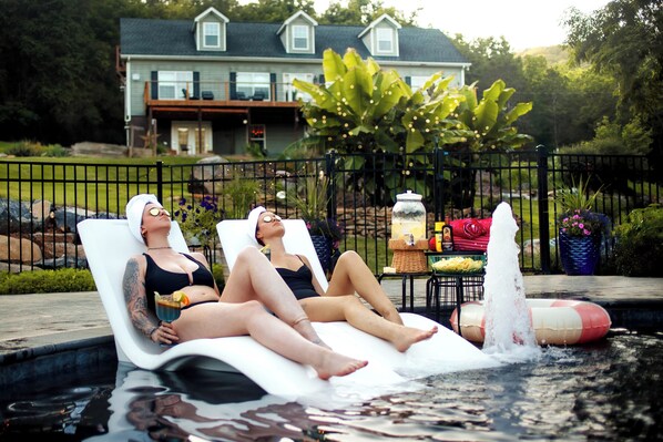 Two individuals relaxing on lounge chairs in a swimming pool with a house and garden in the background.