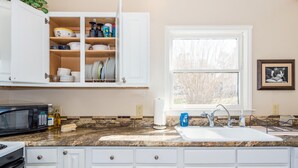 A kitchen with white cabinets, a granite countertop, a built-in microwave, a sink, and a window with a view of bare trees. - Sunset Cottage Blocks from Downtown (Brevard)