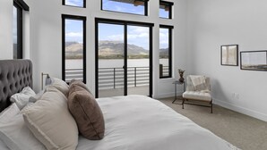 Quarto com uma cama grande, uma cadeira e vista para um lago através de janelas amplas.