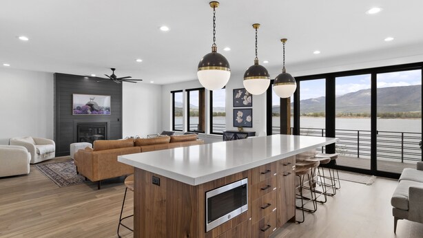 Cozinha moderna com ilha central, armários de madeira e micro-ondas embutido. Há uma área de estar com sofá e cadeiras, e janelas amplas com vista para o exterior.