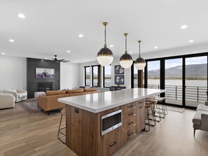 Een moderne keuken met een centraal eiland, houten kasten en een ingebouwde magnetron. Er is een zithoek met een bank en stoelen, en grote ramen met uitzicht op de omgeving.