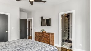 Un dormitorio con cama, cómoda de madera, ventilador de techo y televisor en la pared.