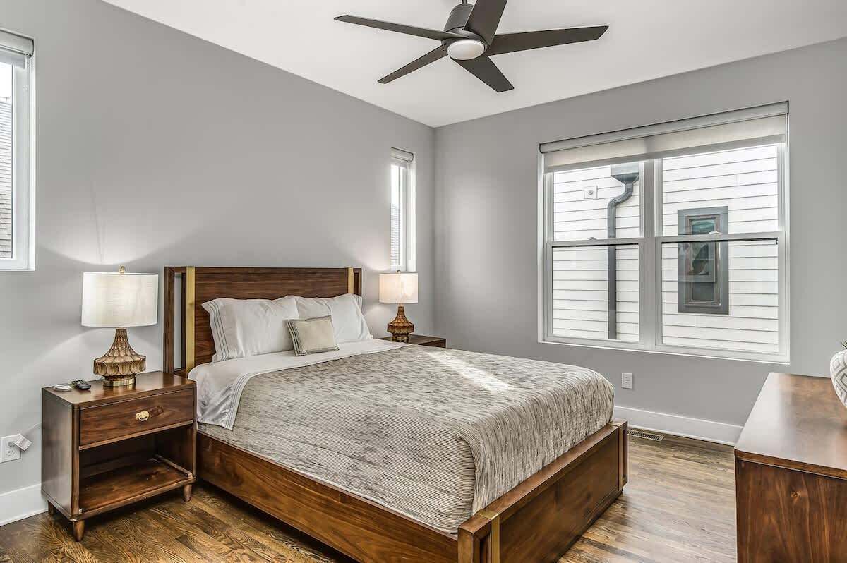 Une chambre à coucher avec un lit en bois, deux tables de chevet avec des lampes, un ventilateur de plafond et une grande fenêtre avec des stores.
