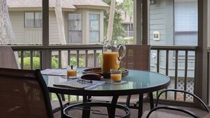 Una mesa de vidrio con una jofaina de jugo de naranja, un vaso de jugo de naranja y una libreta en un porche con vistas a casas.