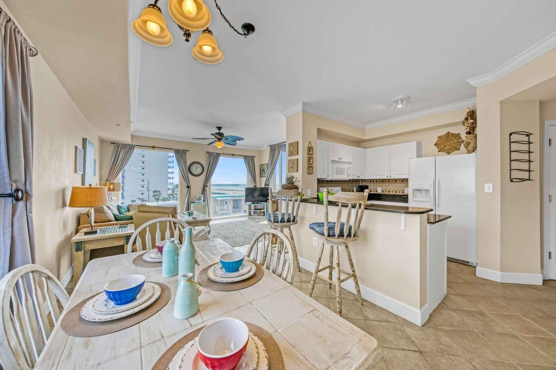 A spacious living area with a dining table set for four, a kitchen with white cabinets, and a view of the city through large windows.