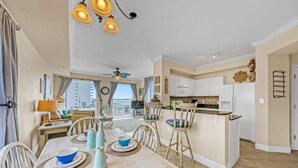 Amplia sala de estar con una mesa de comedor preparada para cuatro, una cocina con armarios blancos y vistas a la ciudad a través de amplios ventanales.