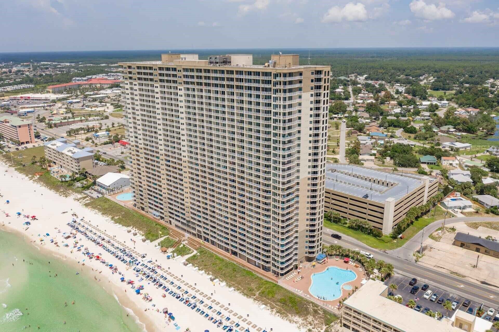 A high-rise hotel with multiple floors, a swimming pool, and a beach area with umbrellas.