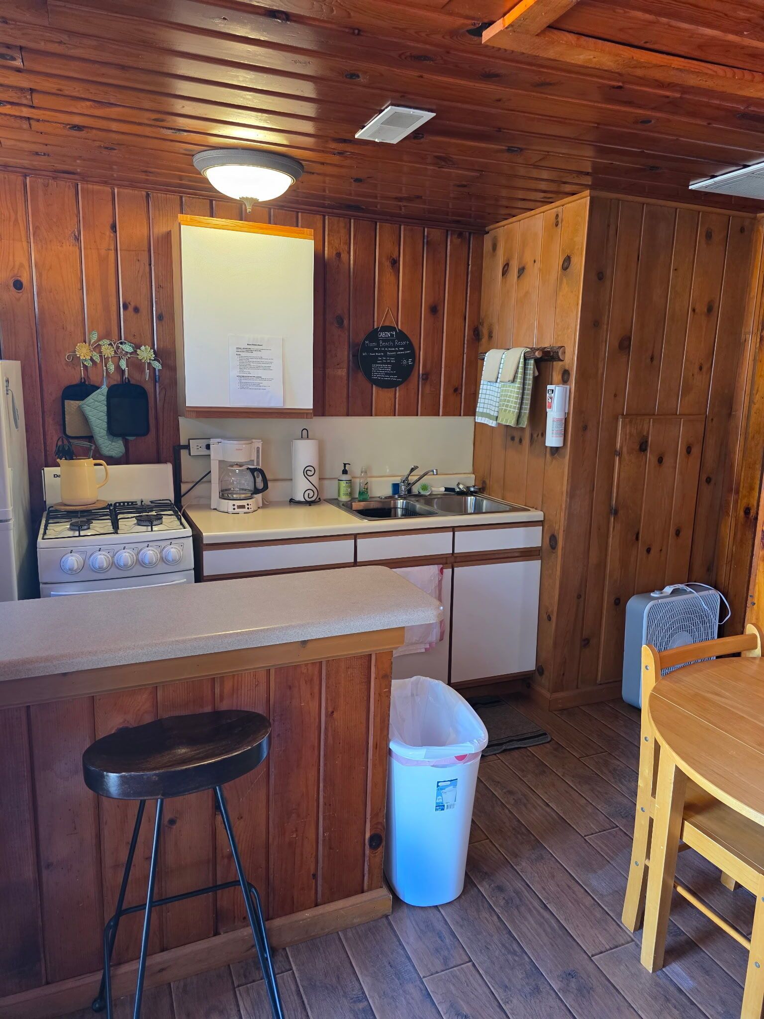 A kitchen with wooden walls, a stove, a sink, a bar counter, and a trash can.