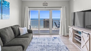 Een woonkamer met een grijze bank, een flatscreen-tv, een groot raam met uitzicht op zee en een balkon met een stoel.