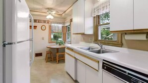 A compact kitchen with white cabinets, a built-in dishwasher, a window with lace curtains, and a small wooden table.