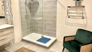 A modern bathroom with a glass-enclosed shower, a wooden vanity, a green chair, and a small plant on a shelf.