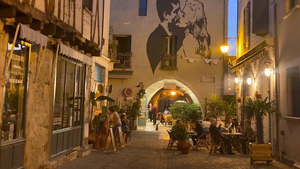Una calle estrecha flanqueada por edificios, un mural de un hombre y un perro, y zonas de estar al aire libre con mesas y sillas.