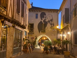 A narrow street with buildings on either side, a mural of a man and a dog, and outdoor seating areas with tables and chairs.