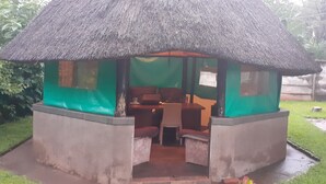 A small, thatched-roof structure with a table and chairs inside, surrounded by greenery. - The Best Green Garden Guest House Harare (Harare)