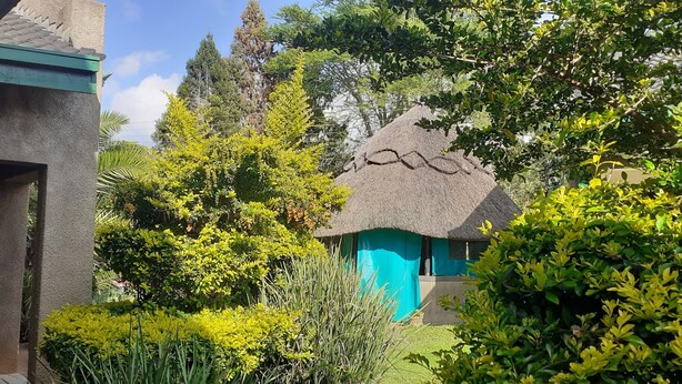 A thatched-roof structure with a turquoise door, surrounded by lush greenery and trees. - The Best Green Garden Guest House Harare (Harare)