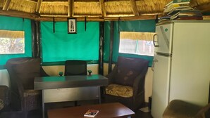 A room with a thatched roof, green walls, a small table, two chairs, a refrigerator, and a window with a view of trees. - The Best Green Garden Guest House Harare (Harare)
