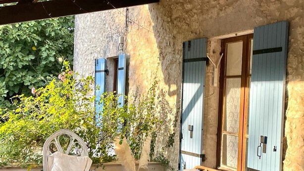 A rustic outdoor seating area with a white wicker chair and a sofa, surrounded by greenery and a stone wall. - Farmhouse Sleeps 11 with Shared Pool (Eymet)