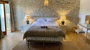 A bedroom with a stone wall, a bed with white linens, two bedside lamps, a bench, and a dresser with a mirror. - Farmhouse Sleeps 11 with Shared Pool (Eymet)