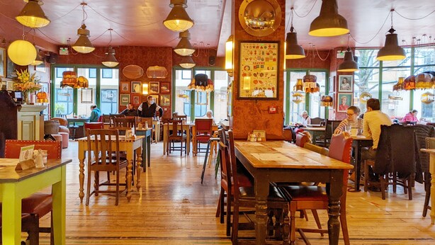 A restaurant with wooden tables and chairs, hanging pendant lights, and large windows.