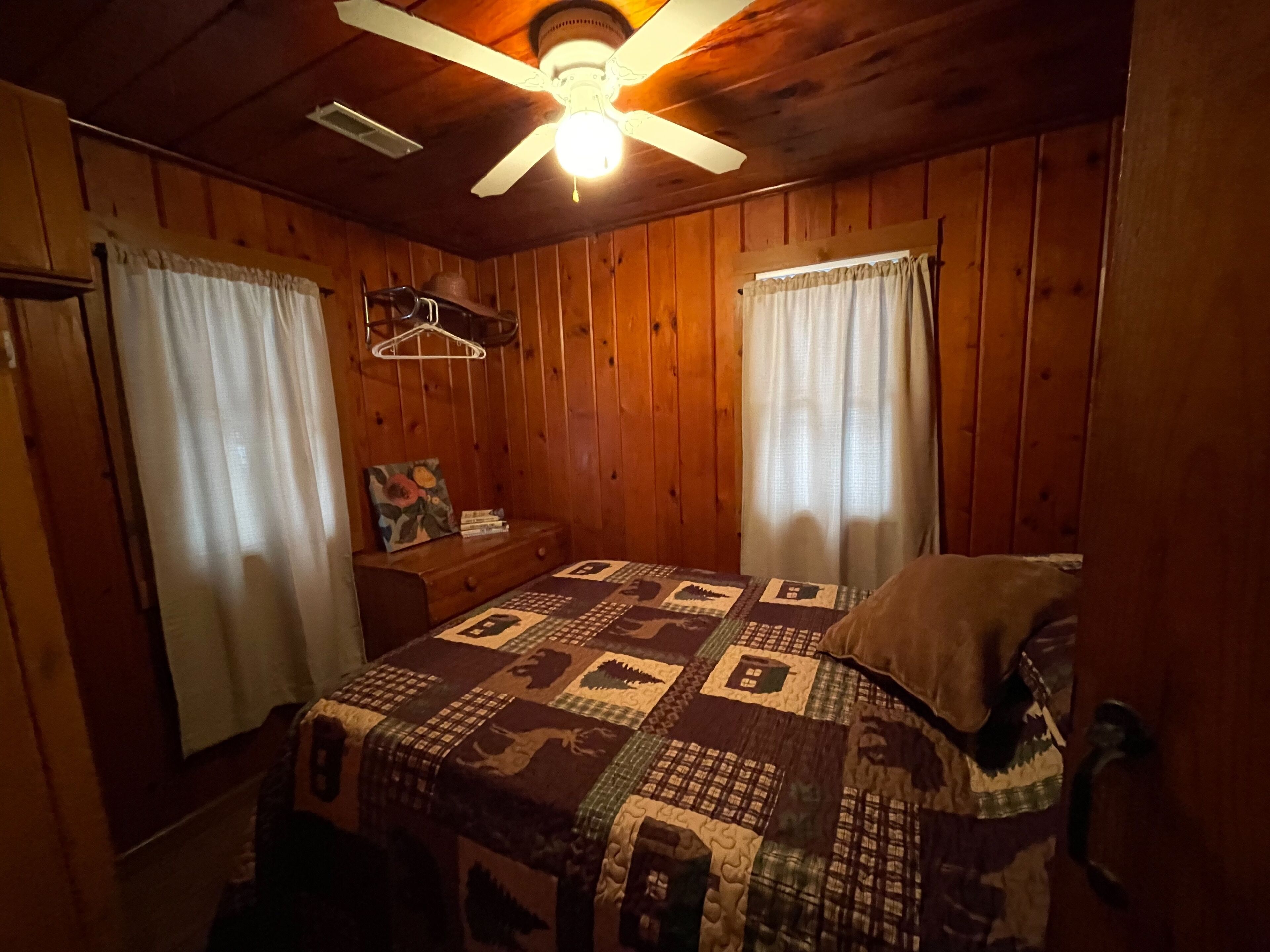 A cozy room with wooden walls, a bed with a quilt, a nightstand, a hanging coat rack, and a ceiling fan.