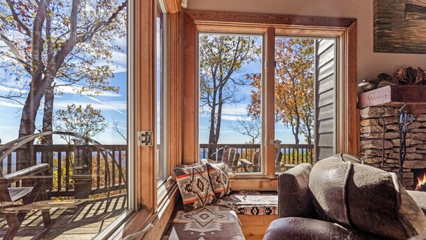 A cozy living room with a large window overlooking a deck and trees, a stone fireplace, and a comfortable seating area. - Enjoy mountain views, cook out on the Weber grill & relax! - Canoe Believe It (Jasper)