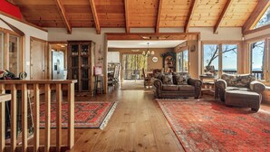 A spacious living room with wooden floors, a high ceiling, and large windows offering a view of the outdoors. - Enjoy mountain views, cook out on the Weber grill & relax! - Canoe Believe It (Jasper)
