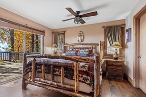 Une chambre à coucher rustique avec un lit en bois, un ventilateur de plafond, une table de chevet et une grande fenêtre donnant sur les arbres aux couleurs automnales.