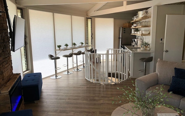 Un salon moderne avec un plancher en bois, un pouf bleu, un canapé gris et une barrière de sécurité blanche pour bébé. Il y a également un coin cuisine avec un réfrigérateur, des étagères et une table à manger avec des chaises.