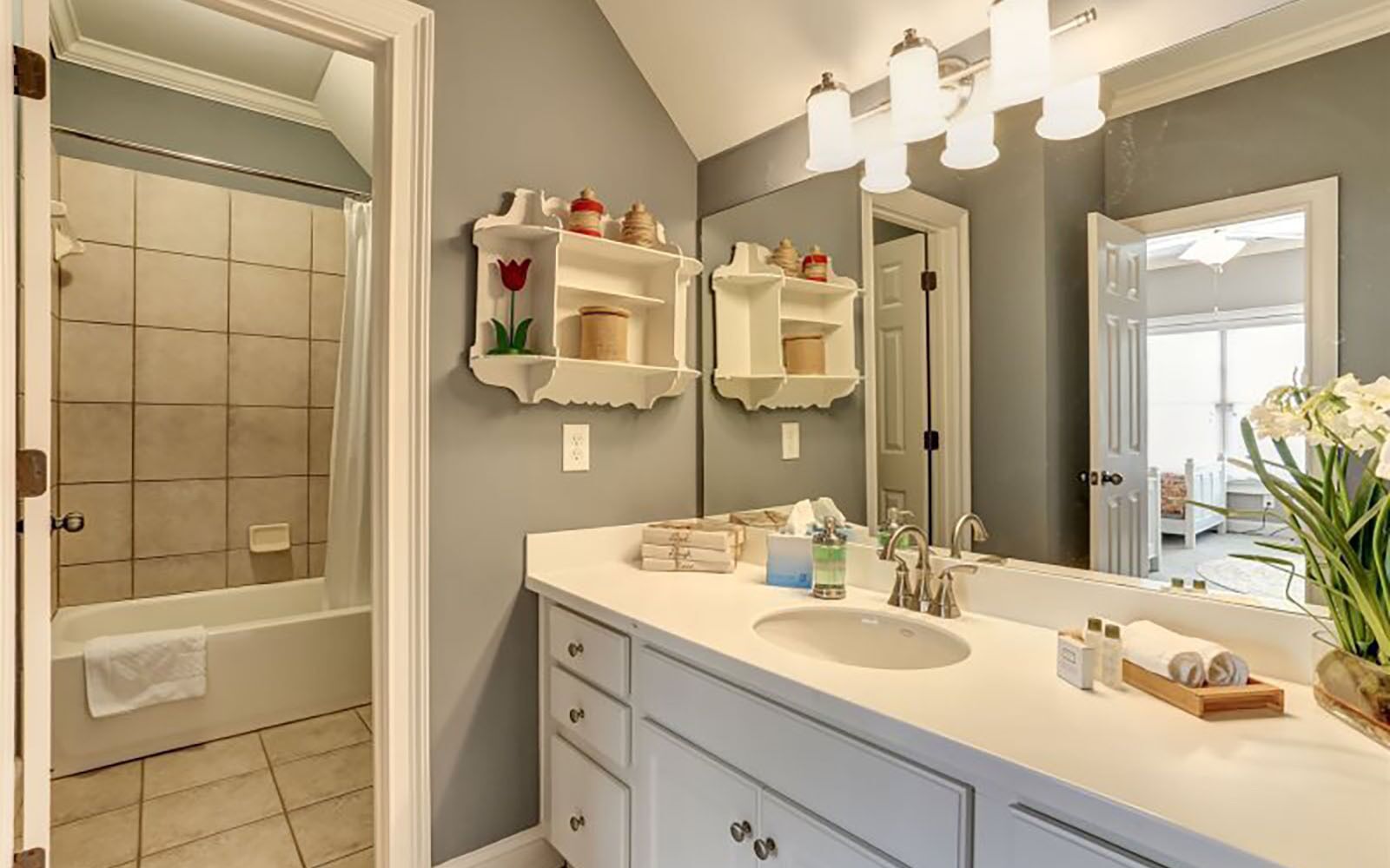 Une salle de bain dotée d’un grand miroir, d’un meuble-lavabo blanc et d’étagères où sont rangés les produits de toilette.