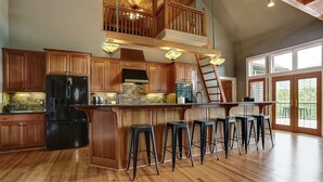 A spacious kitchen with wooden cabinets, a black refrigerator, a large island with bar stools, and a loft area with a wooden staircase. - Fireside Sips, Soothing Hot Tub Soaks, Hiking Trails, Lakeside Dining & Scenic Golf - Field of Stars (Jasper)