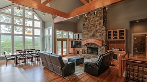 A spacious living room with a stone fireplace, wooden beams, and large windows offering a view of the outdoors. - Fireside Sips, Soothing Hot Tub Soaks, Hiking Trails, Lakeside Dining & Scenic Golf - Field of Stars (Jasper)