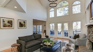 A spacious living room with a dark gray sofa, a coffee table with a plant, a stone fireplace, and a dining area visible through glass doors. - Hillside Manor - Luxury Hot Tub + Fireplace + Pool Table + & Wet Bar (Jasper)