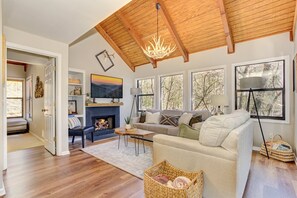 A spacious living room with a fireplace, a sofa, a coffee table, and a large window with a view of trees. - Laughing Fox - Treehouse Vibes, Firelit Moments, Movie Nights, Golf & Trails (Dawsonville)