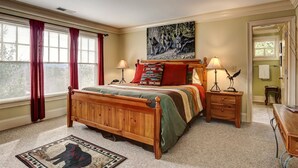 Une chambre à coucher avec un lit en bois, deux tables de chevet avec des lampes, un tableau au mur et une vue sur l’extérieur par une fenêtre ornée de rideaux rouges.