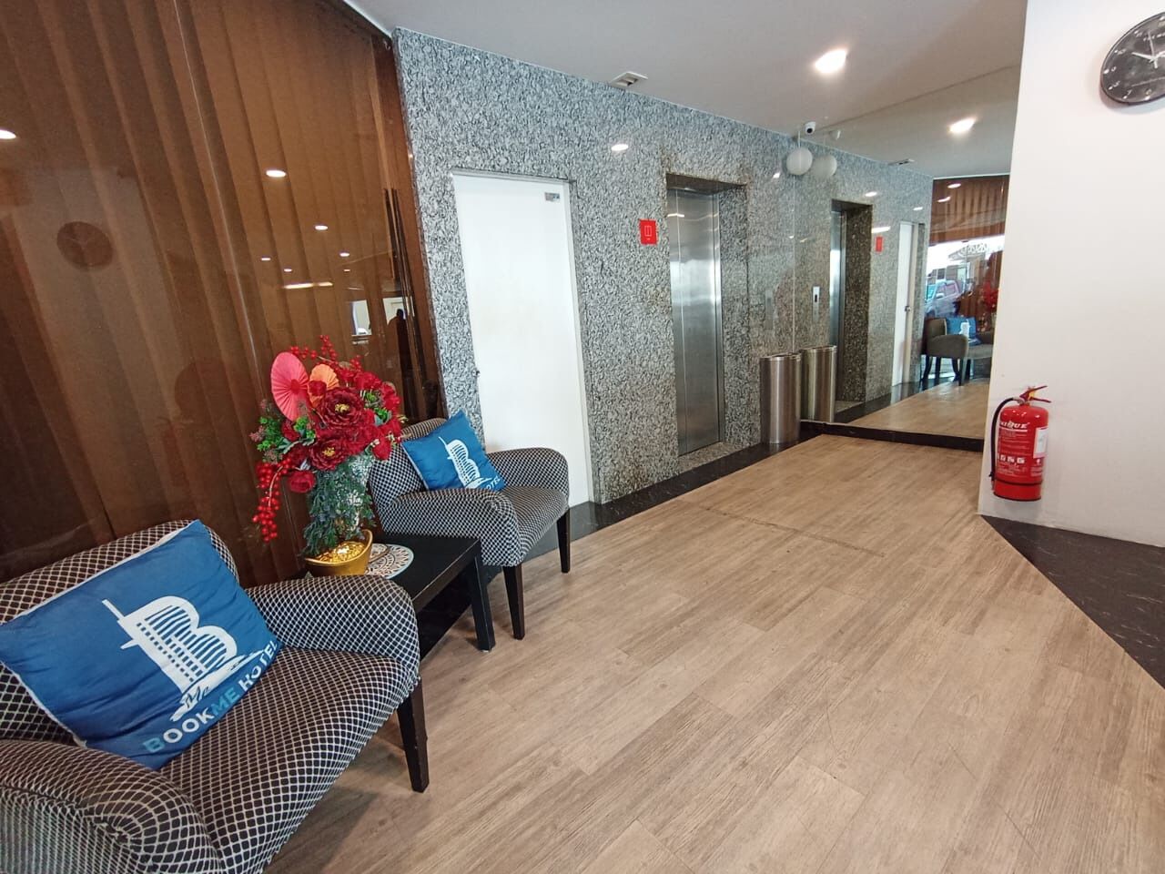 A hotel reception area with a check-in desk, a seating area with patterned sofas, a decorative plant, and a fire extinguisher.