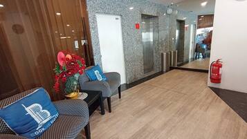 A hotel reception area with a check-in desk, a seating area with patterned sofas, a decorative plant, and a fire extinguisher.
