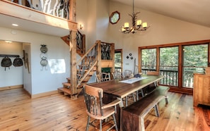 A rustic wooden interior with a large dining table, wooden chairs, a staircase with exposed beams, and large windows offering a view of the outdoors.