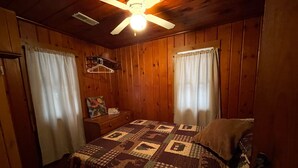 Une pièce chaleureuse aux murs en bois, avec un lit recouvert d’une courtepointe, une table de chevet, un porte-manteau suspendu et un ventilateur de plafond.