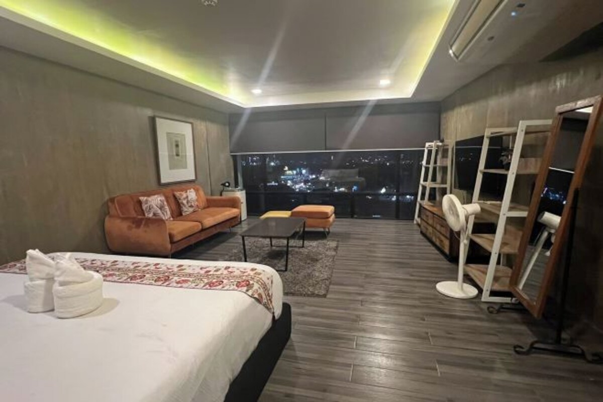 Habitación de hotel moderna con una cama grande, un sofá, una mesita de café y vistas a la ciudad de noche.